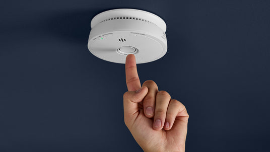 A finger pressing the button on a smoke alarm mounted on the ceiling.