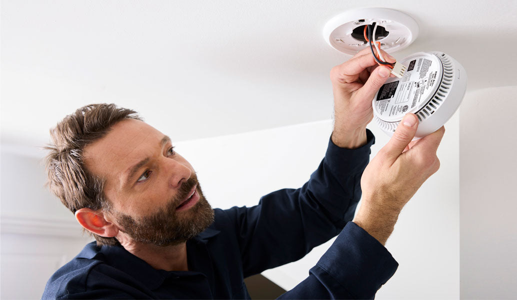 A man in a navy blue shirt installs a white round smoke detector by wiring it to a ceiling fixture in a white room.
